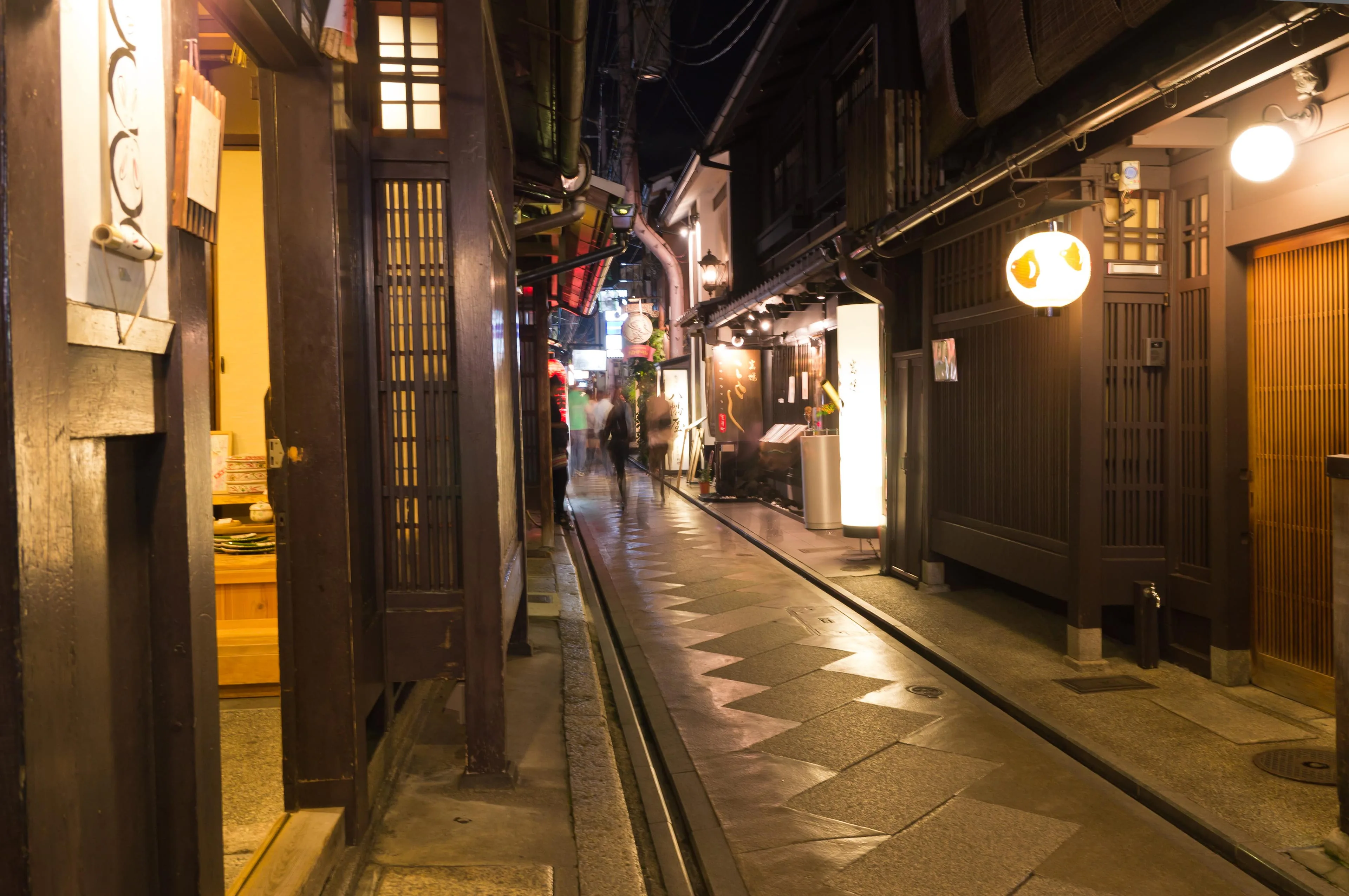 A narrow Kyoto lane in the evening: wooden fronts, lit lanterns, wet stone underfoot